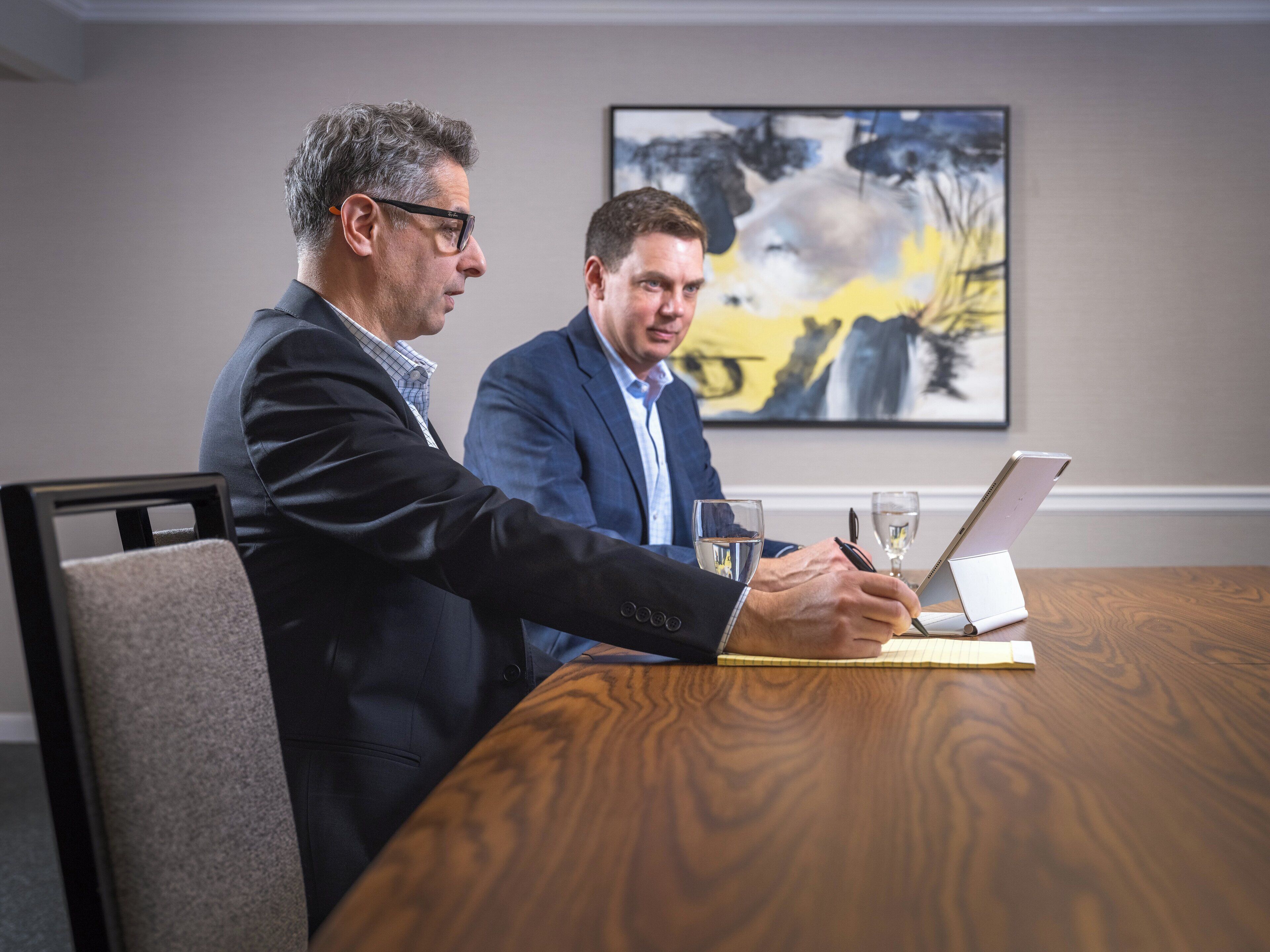 Two men in a meeting room with a wooden table, a laptop, and a glass of water.