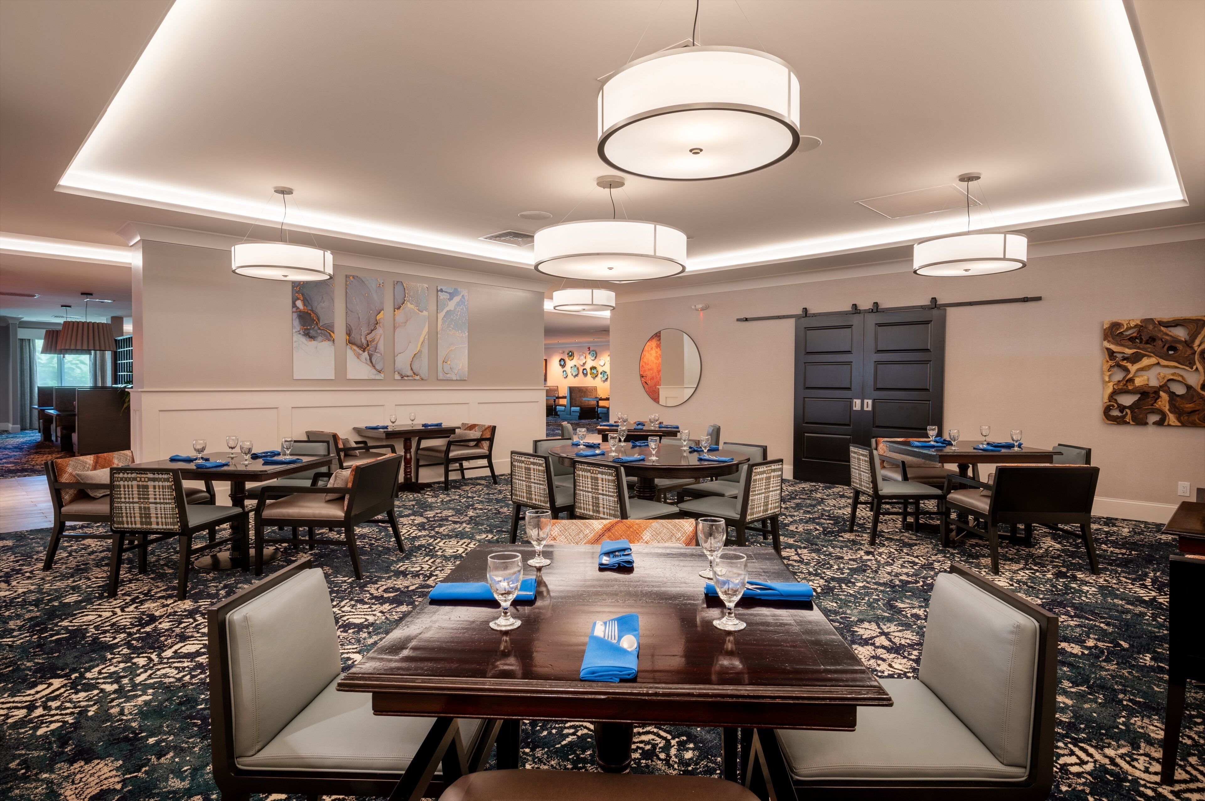 Un comedor de hotel con varias mesas y sillas, iluminación colgante moderna y una alfombra estampada.