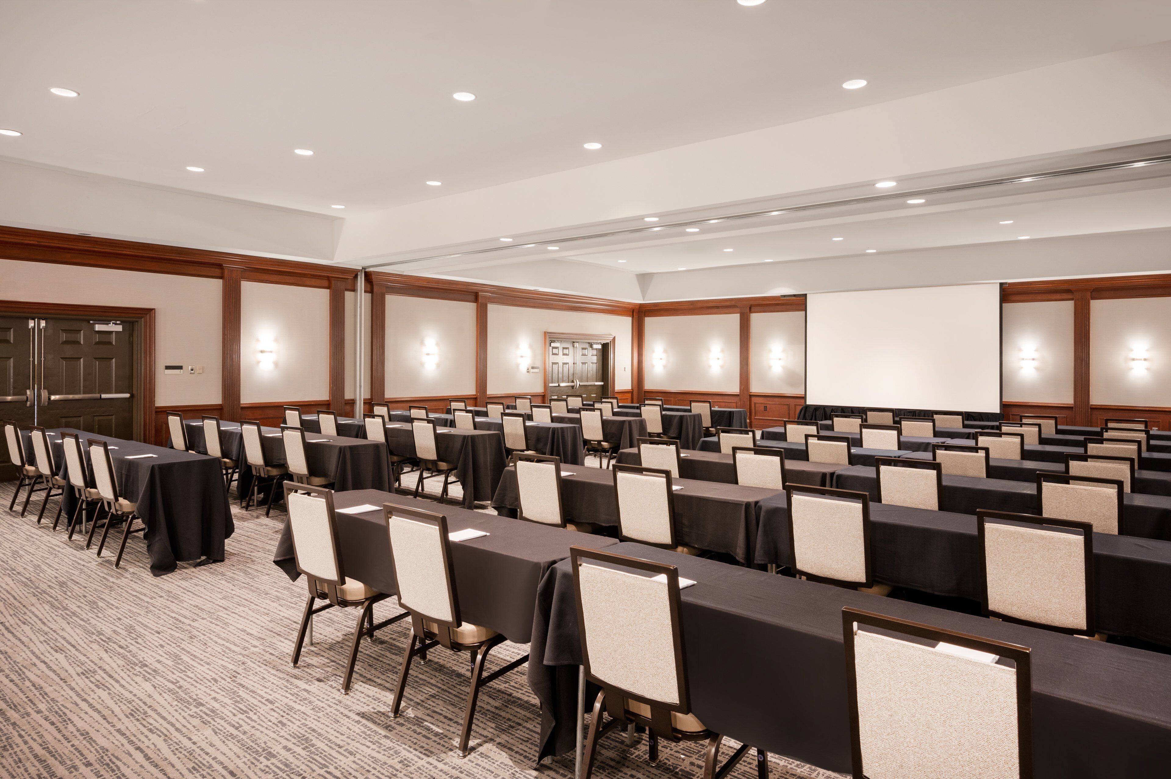 A large conference room with rows of tables and chairs, equipped with overhead lighting and a projection screen.