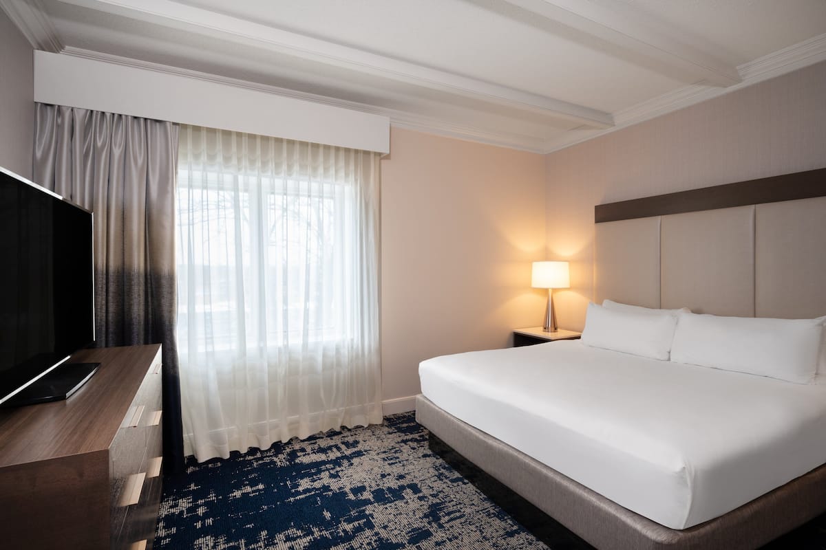 Een hotelkamer met een groot bed, een televisie op een houten bureau, een lamp en een raam met dunne gordijnen.