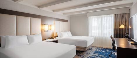 A hotel room with two beds, a desk, a chair, a lamp, and a large window with curtains.