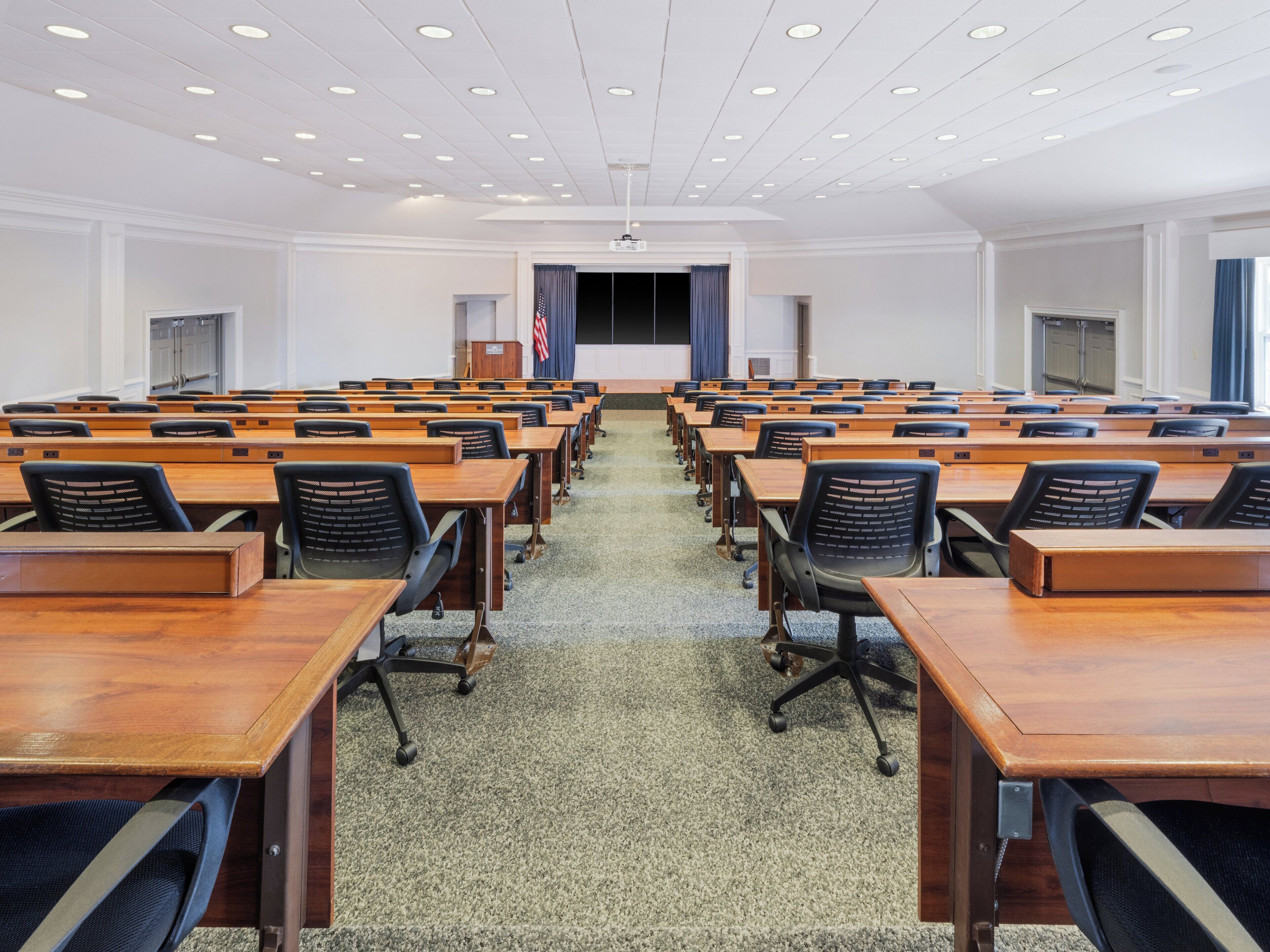 Une salle de conférence avec des rangées de tables en bois et des chaises de bureau noires, un sol recouvert de moquette et un plafond blanc doté d’un éclairage encastré.