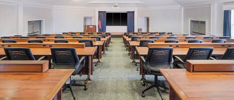 A conference room with rows of wooden tables and black office chairs, a carpeted floor, and a white ceiling with recessed lighting.