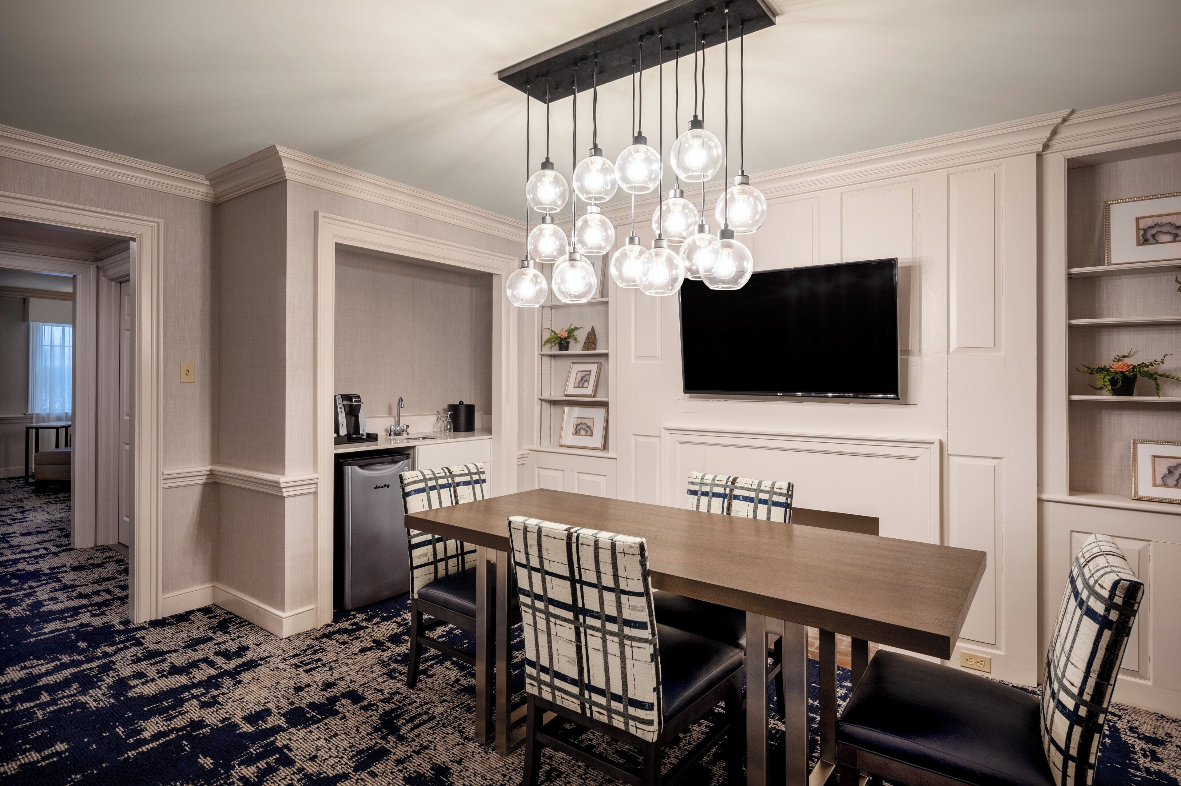 A dining area with a wooden table, six chairs, a television mounted on the wall, and a kitchen area with a coffee machine.