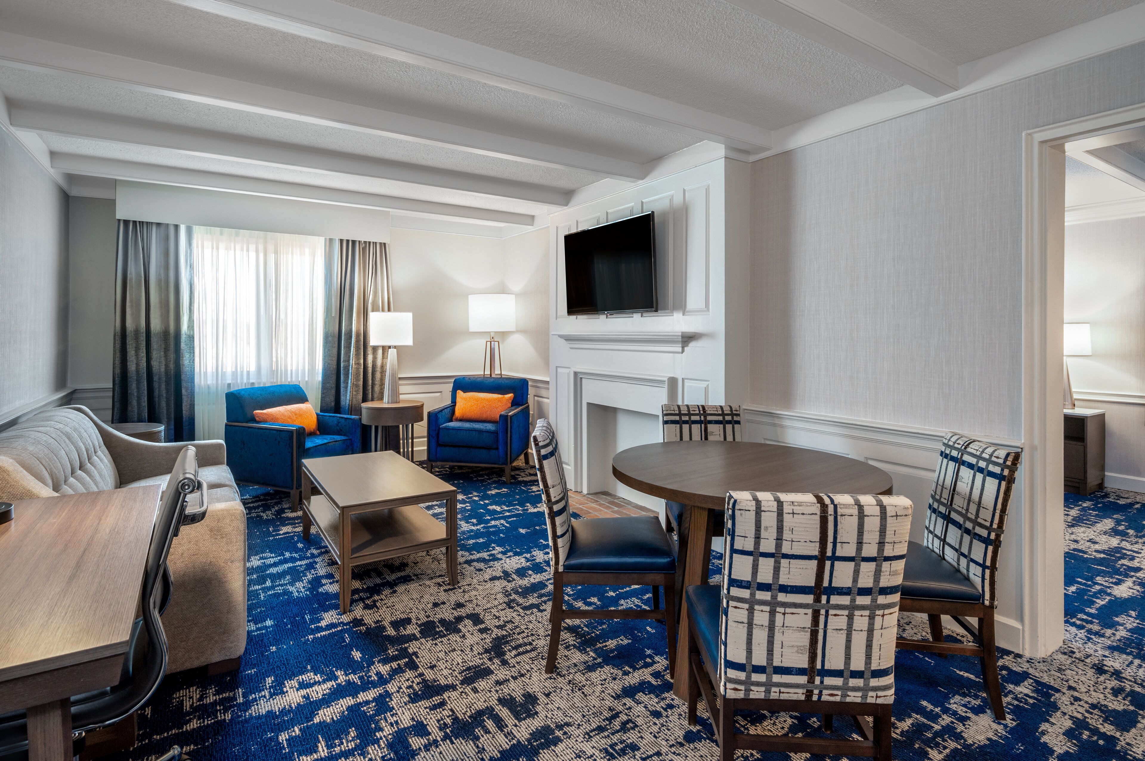 Hotelkamer met een zithoek, een eettafel, blauwe en beige stoelen, een televisie boven een open haard, en een kamerbreed tapijt met dessin.