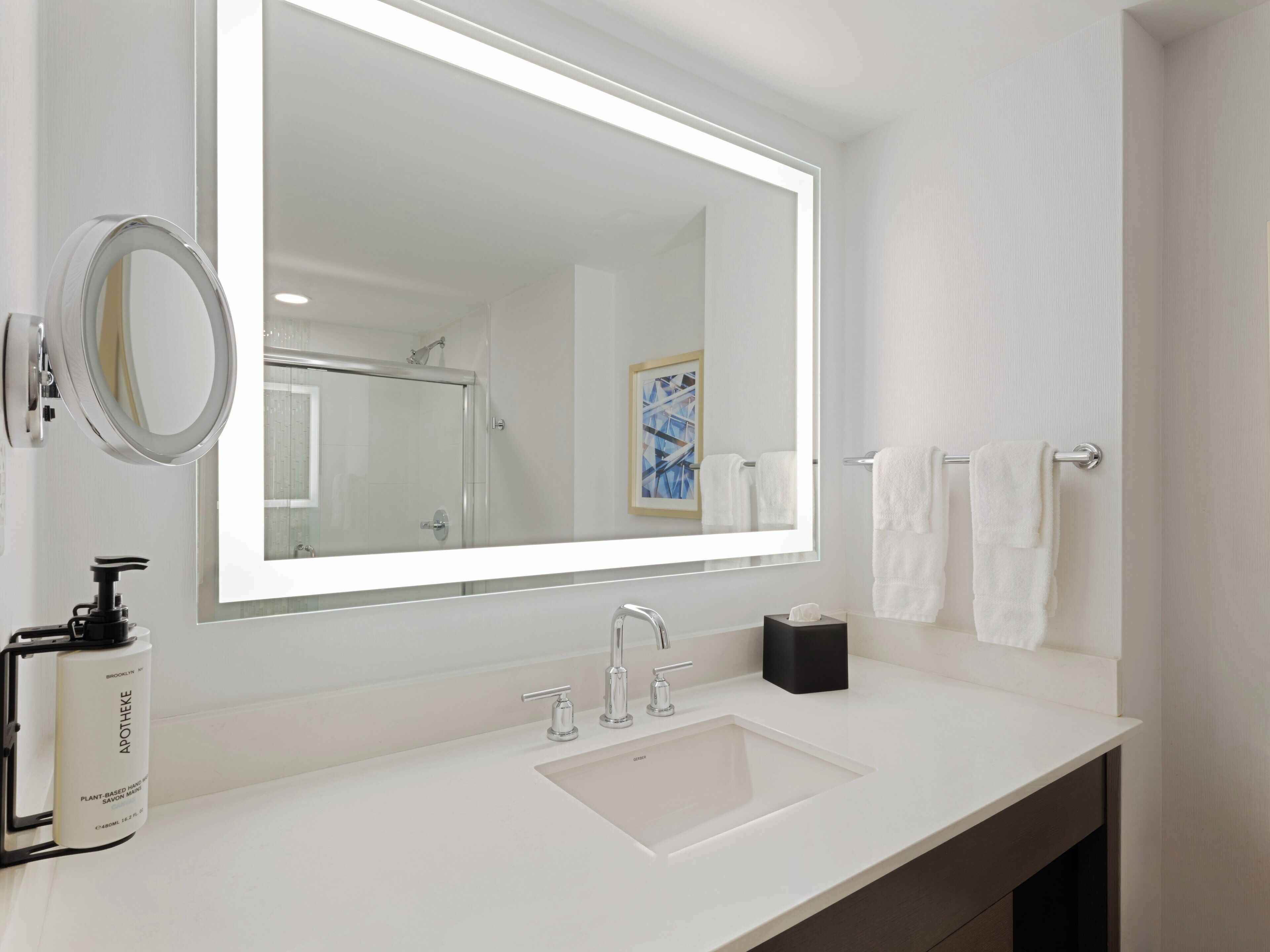 A modern bathroom with a large mirror, a sink, a handheld soap dispenser, and a towel rack.