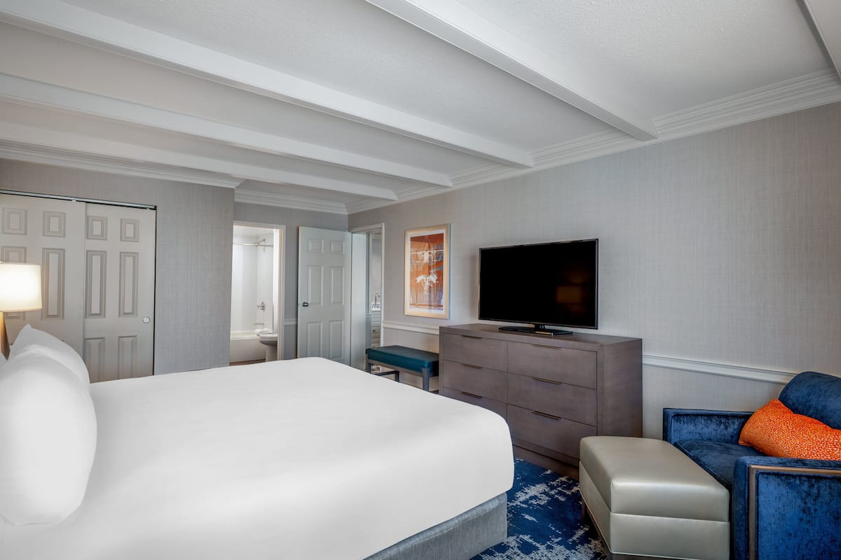 Een hotelkamer met een groot bed, een televisie, een dressoir en een zithoek met een blauwe stoel en een oranje kussen.