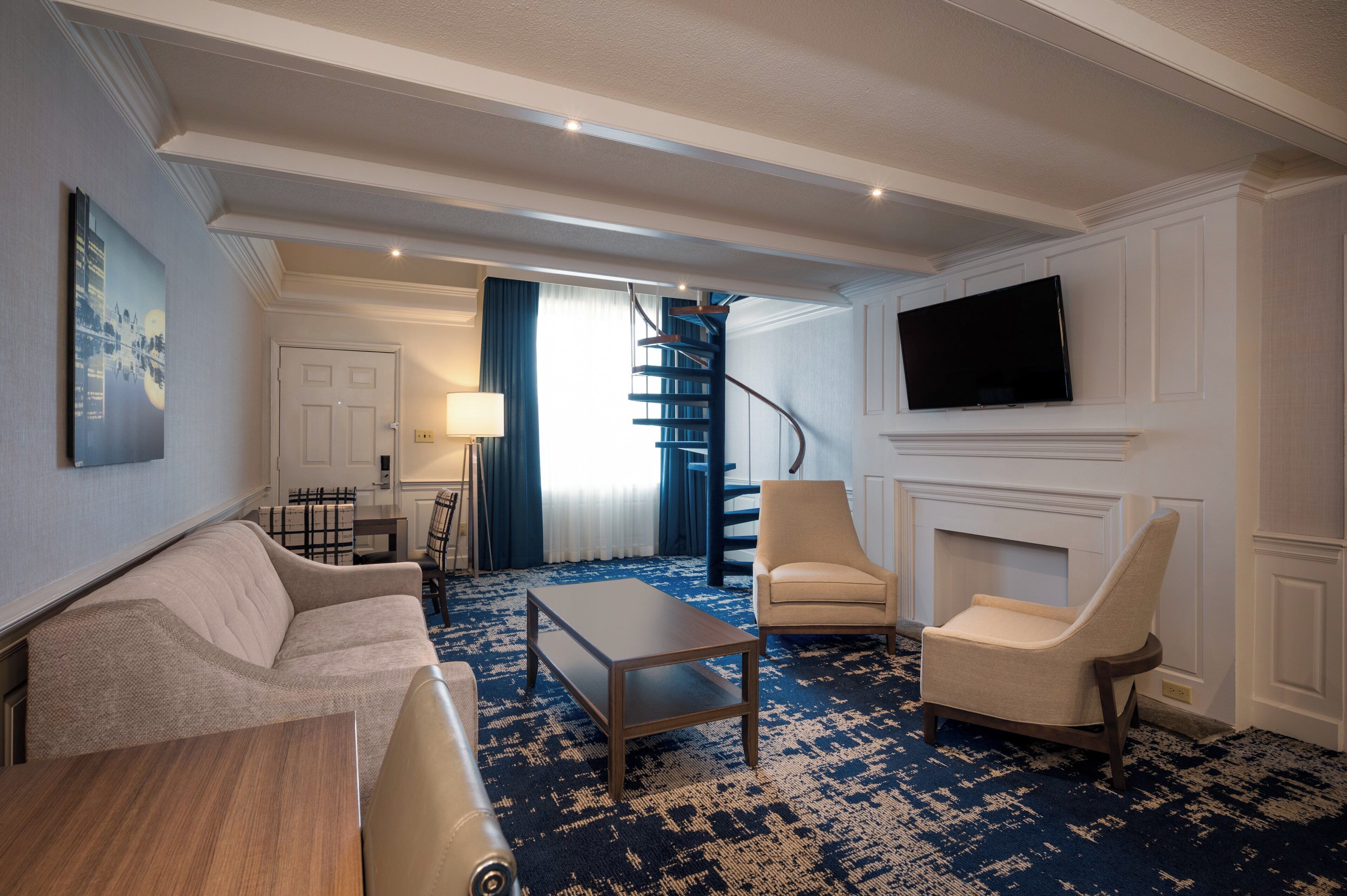 A hotel room with a sofa, armchairs, a coffee table, a flat-screen TV, a fireplace, and a staircase.