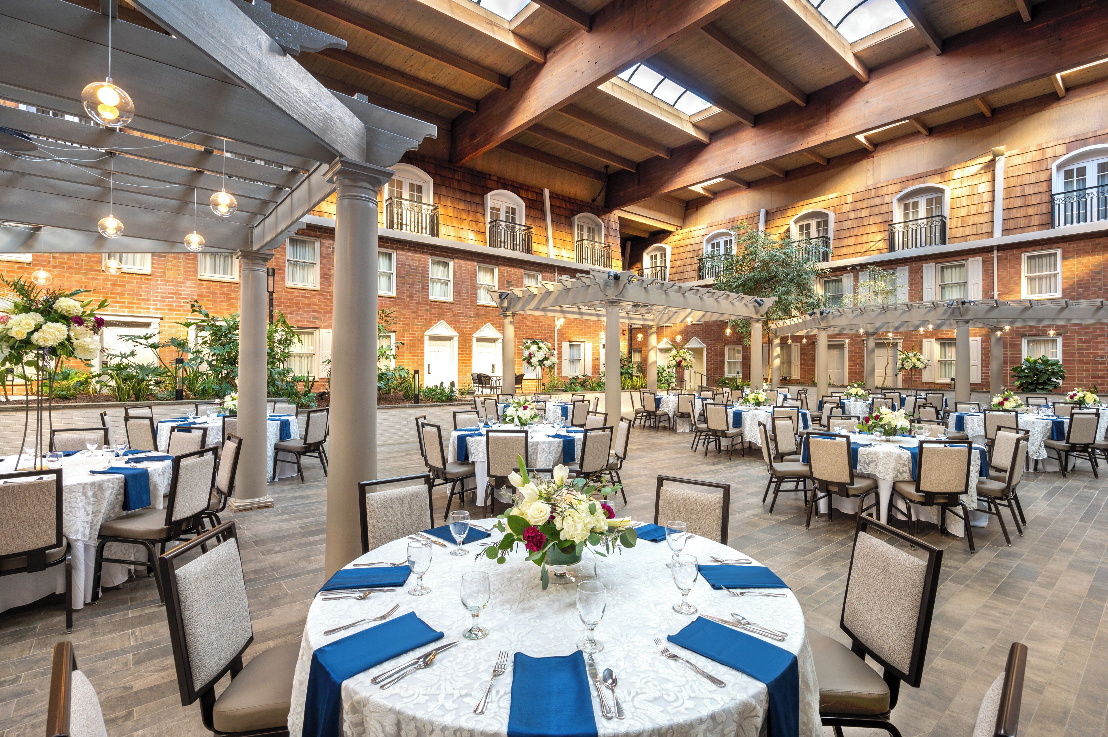 A spacious outdoor dining area with multiple round tables set for guests, surrounded by brick walls and wooden beams.