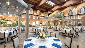 A spacious outdoor dining area with multiple round tables set for guests, surrounded by brick walls and wooden beams.