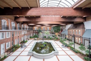 Une cour intérieure avec une fontaine centrale, entourée de bâtiments en briques dotés de fenêtres en arc de cercle et d’espaces aménagés pour s’asseoir à l’extérieur.