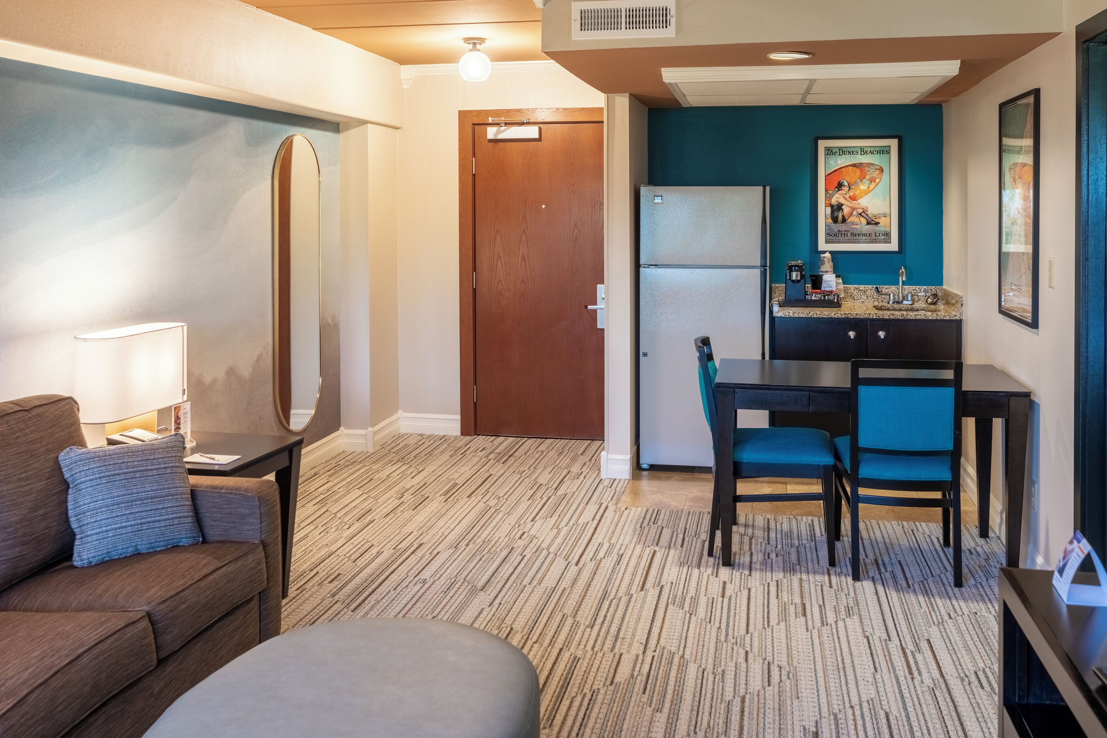Une chambre d’hôtel comprenant un canapé, une petite table avec une lampe, une kitchenette avec un réfrigérateur, un coin repas avec une table et des chaises, et un grand miroir.