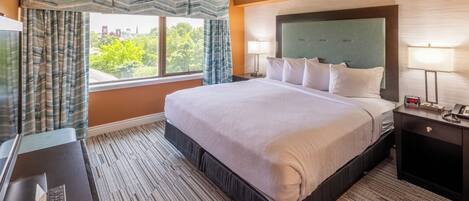 Habitación de hotel con una cama grande, un escritorio con computadora, una ventana con cortinas y una mesita de noche con lámpara.