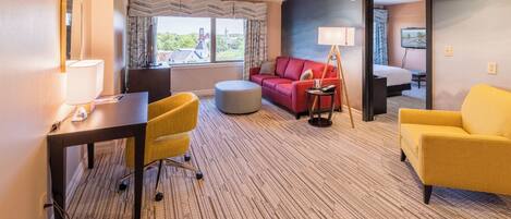 Habitación de hotel con escritorio, dos sillones amarillos, un sofá rojo, un reposapiés redondo, un espejo grande y una televisión.