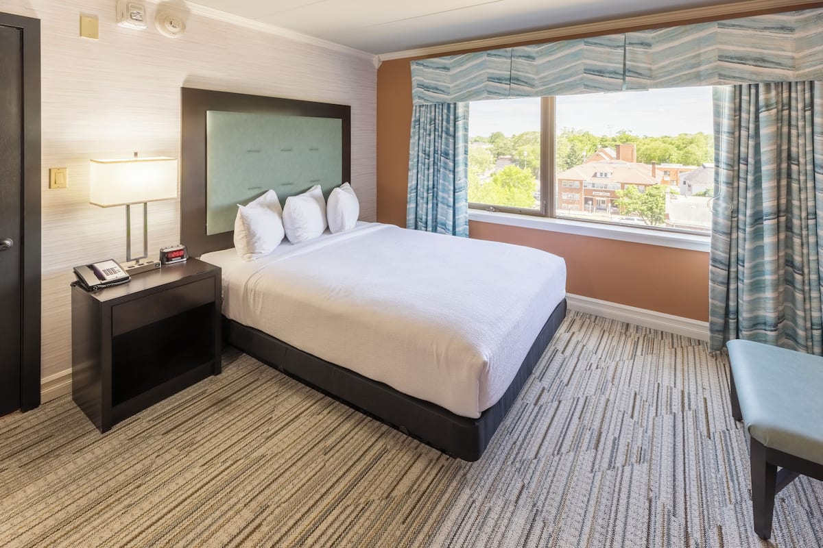 A hotel room with a large bed, a bedside table with a lamp, a phone, and a window with curtains.