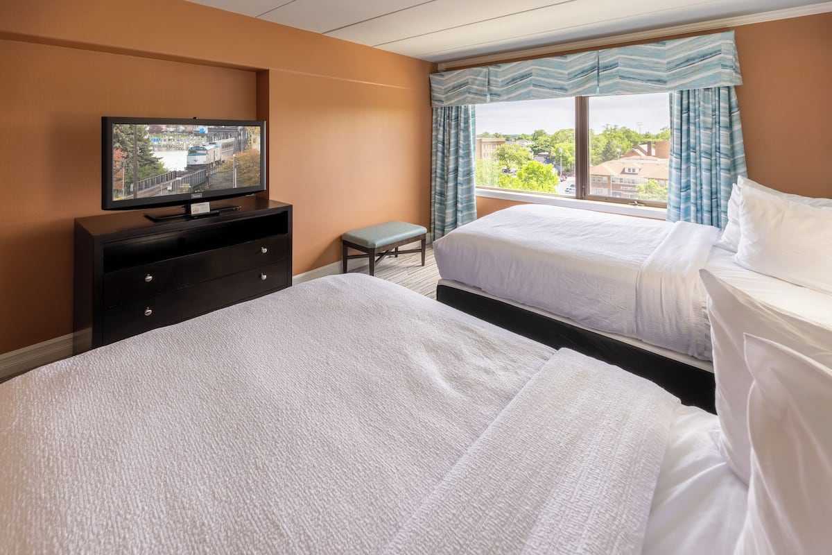 A hotel room with two beds, a television on a dresser, a bench, and a window with curtains.