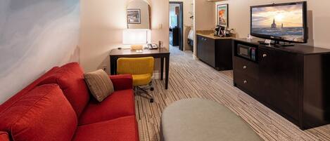 Una habitación de hotel con sofá rojo, sillón amarillo, reposapiés gris, televisor sobre un mueble bajo y escritorio con lámpara.