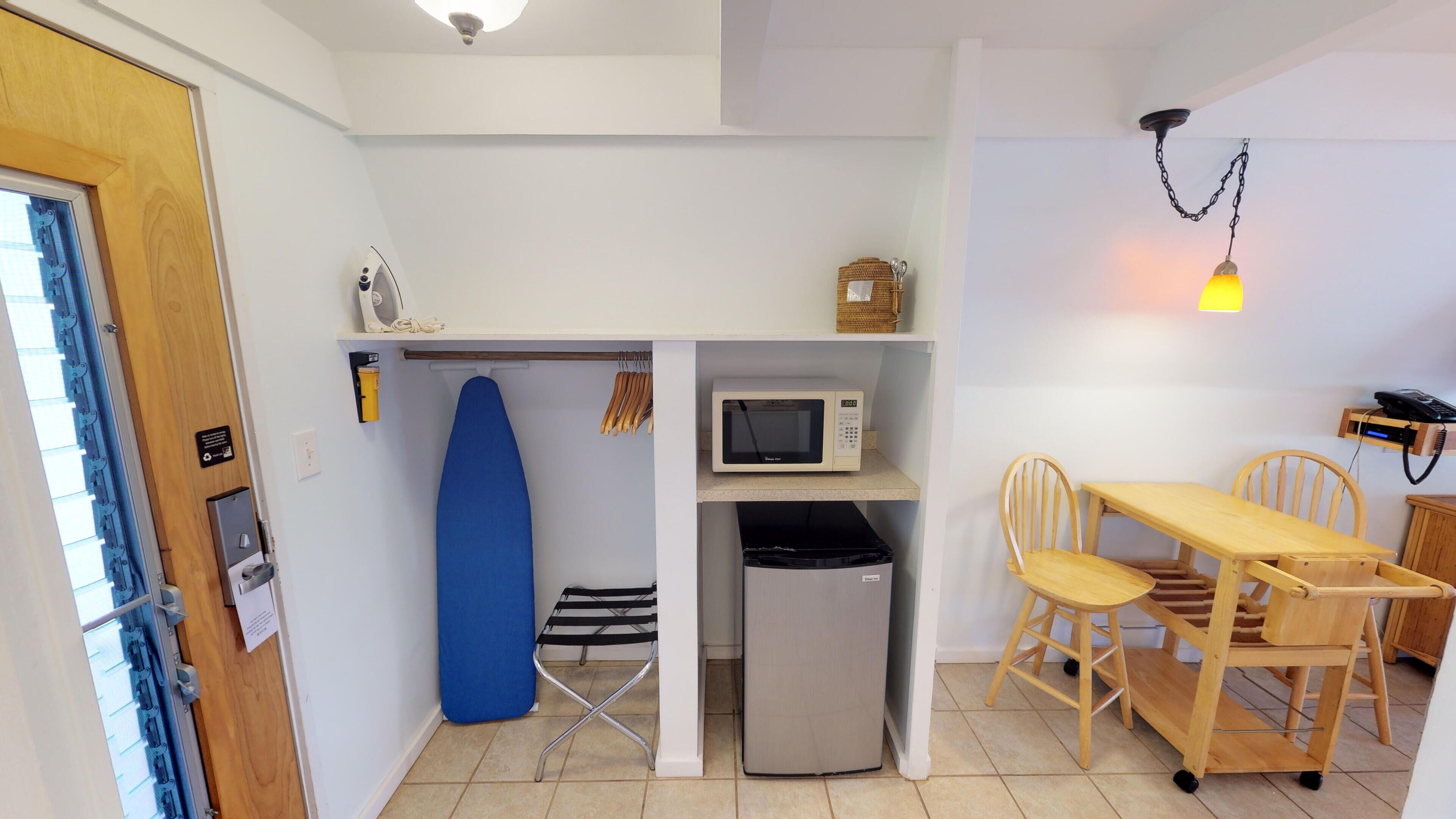 A compact kitchen with a microwave, ironing board, and a small trash bin.