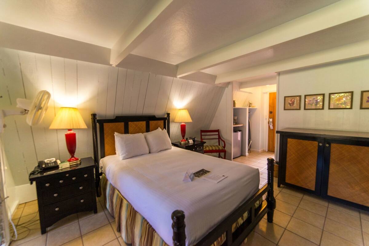 Ein Schlafzimmer mit Bett, Nachttischlampen, einem Nachttisch, einem Sessel, einem Schreibtisch und einer kleinen Küchenzeile.