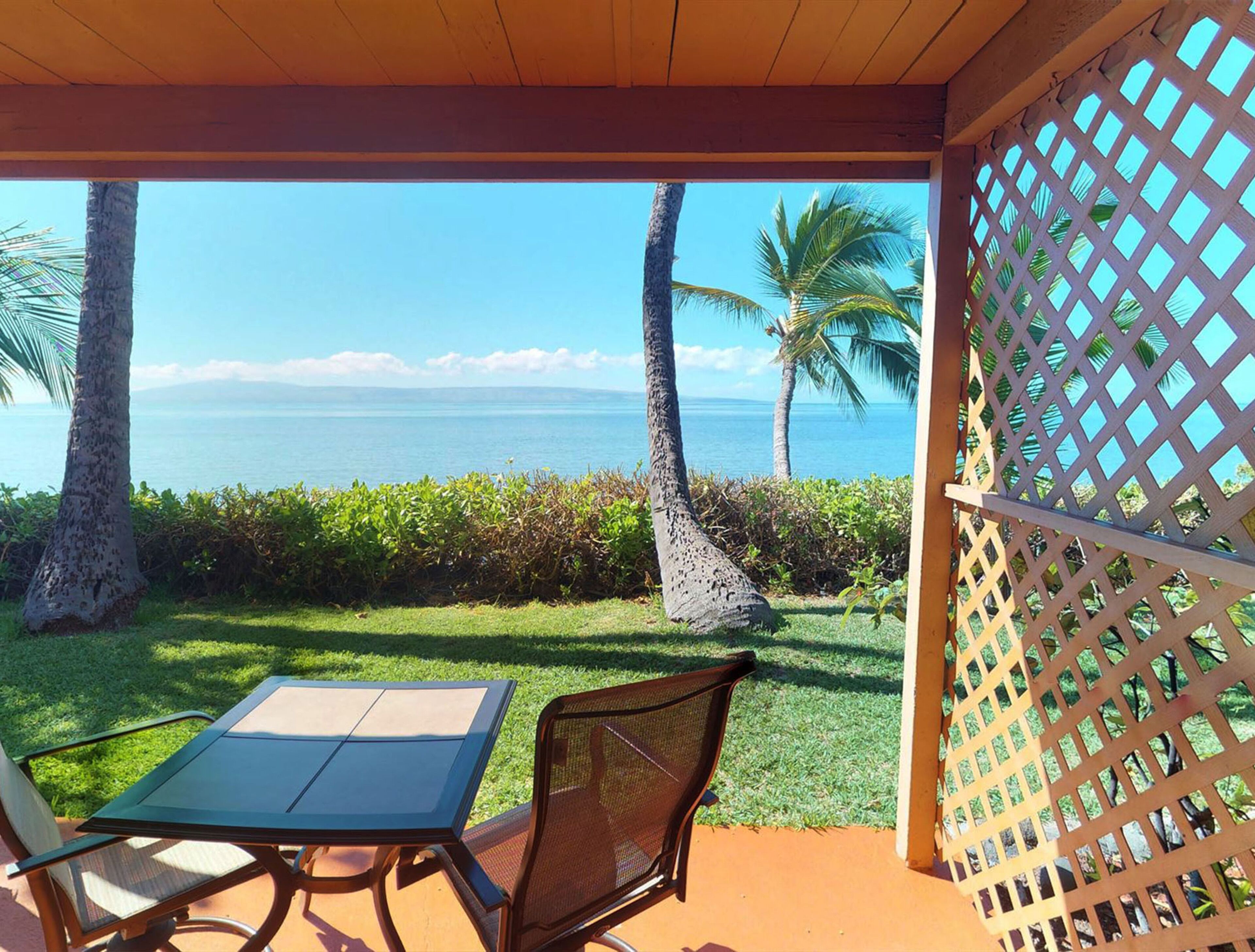 Un espacio de estar techado al aire libre con una mesa y sillas, con vista a una playa con palmeras y el océano.