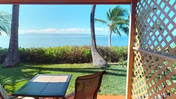 Un espacio de estar techado al aire libre con una mesa y sillas, con vista a una playa con palmeras y el océano.
