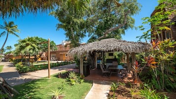 A thatched-roof outdoor seating area with a table and chairs, surrounded by lush greenery and palm trees.