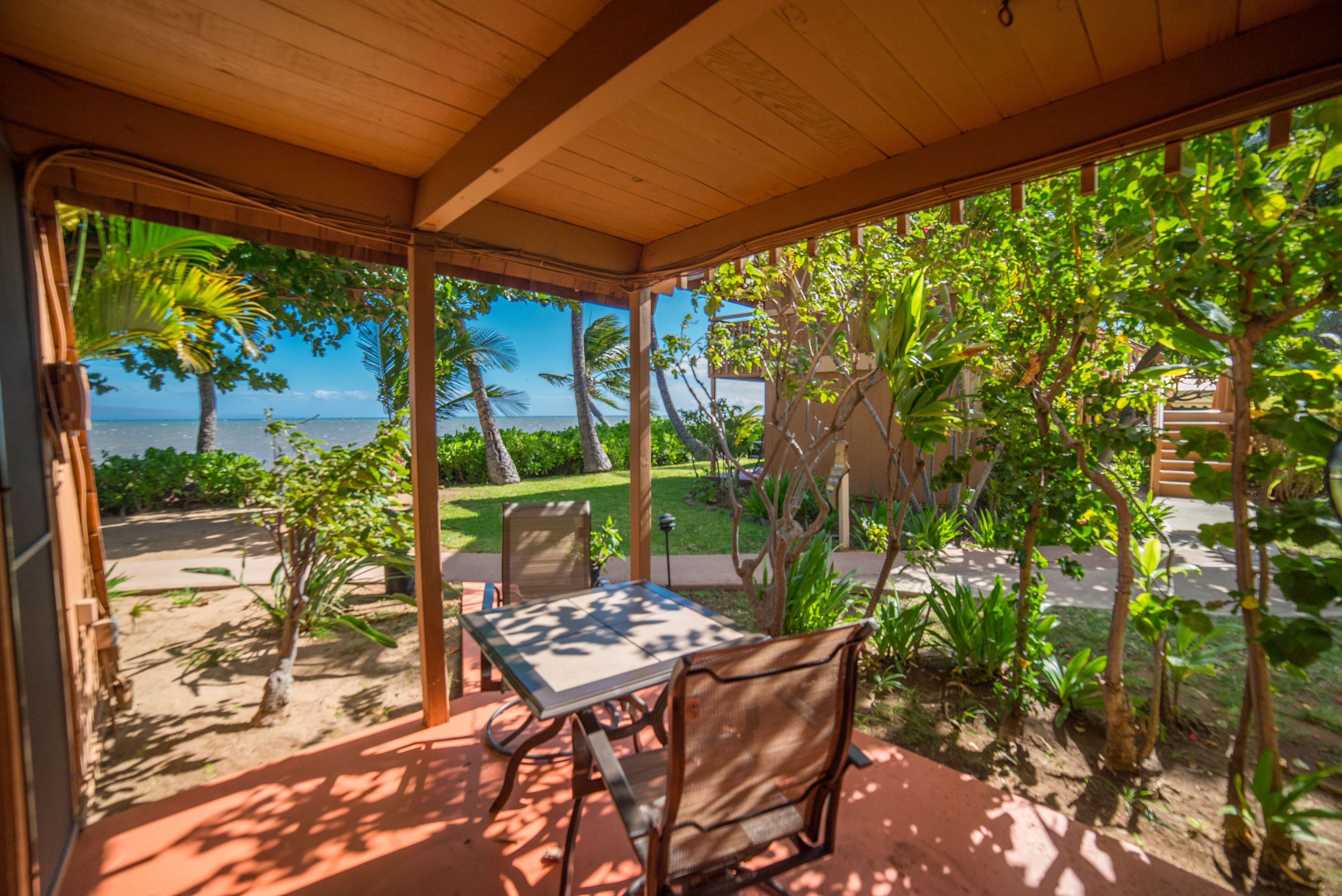 A covered outdoor seating area with a table and chairs, overlooking a garden and a view of the sea.
