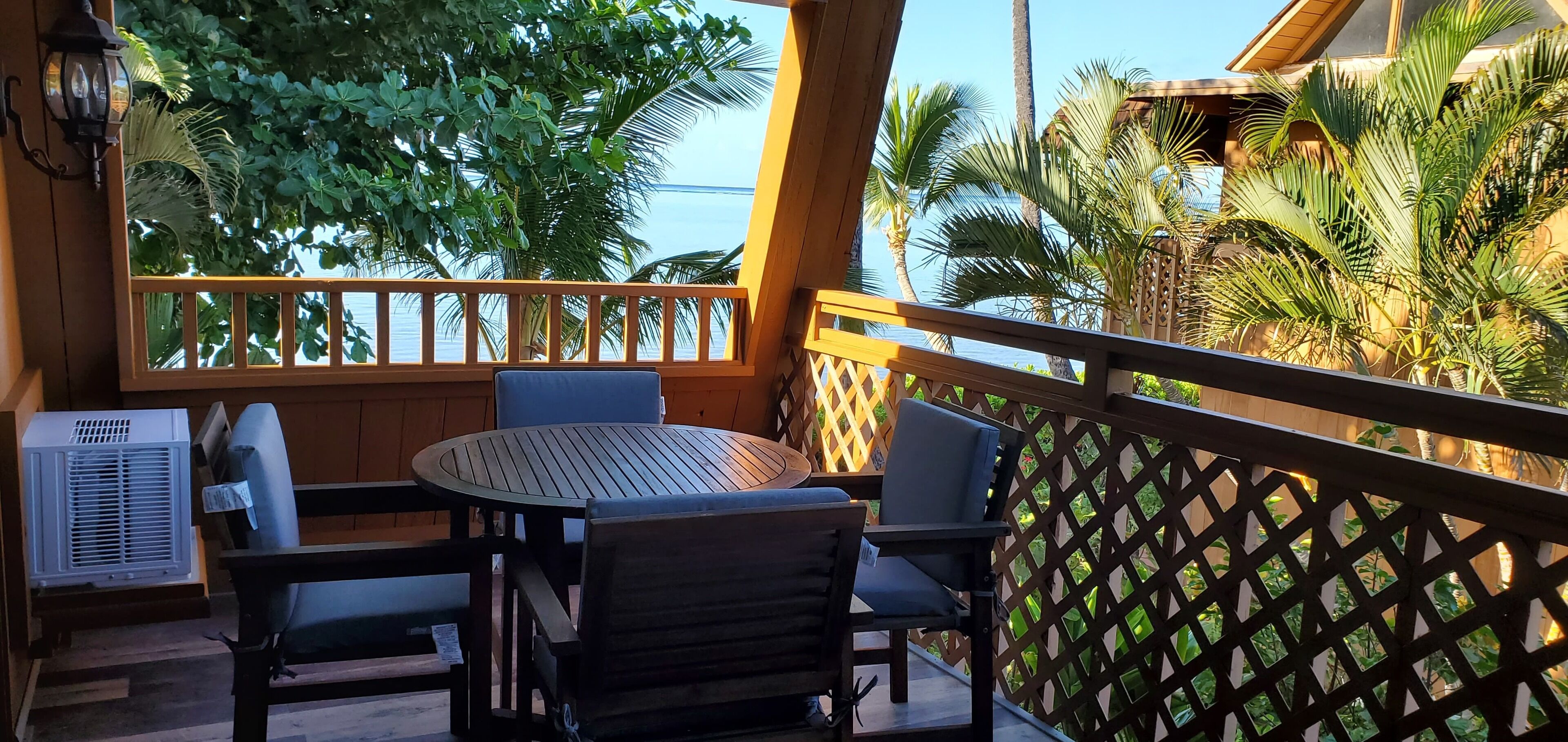 A balcony with a round table and chairs, a wooden railing, and a view of palm trees and the sea.