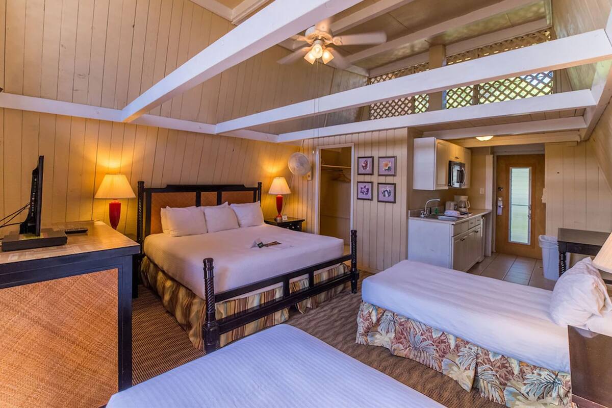 Ein Hotelzimmer mit einem großen Bett, einem kleineren Bett, einem Schreibtisch mit Lampe, einem Deckenventilator und einer Küchenzeile.