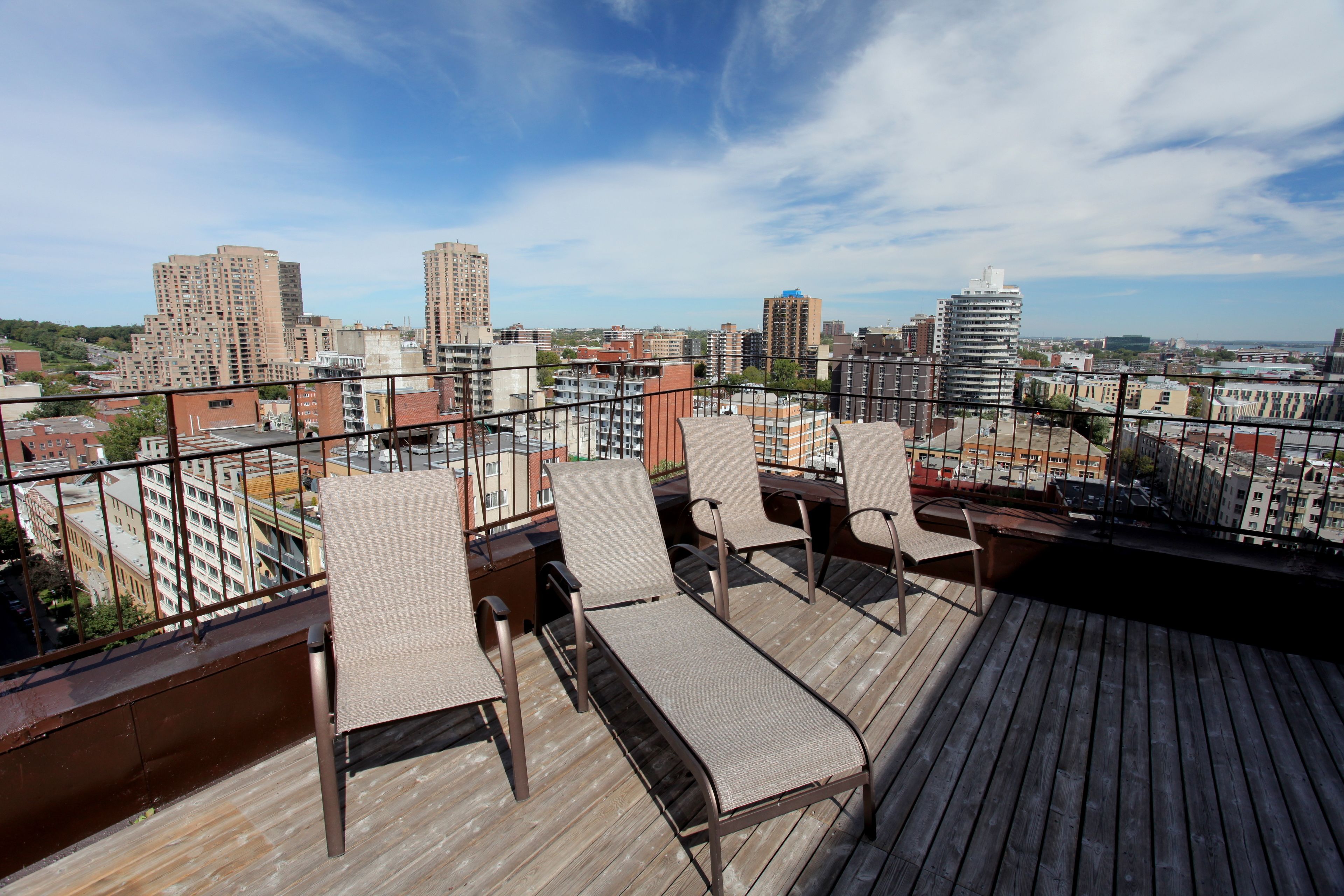A rooftop terrace with four lounge chairs and a wooden deck, overlooking a cityscape with various buildings.