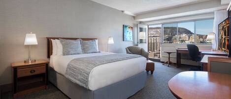 A hotel room with a large bed, a desk, a chair, and a view of the city through large windows.