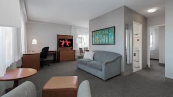 Een hotelkamer met een grijze bank, een houten bureau, een televisie, een schilderij en een kitchenette.