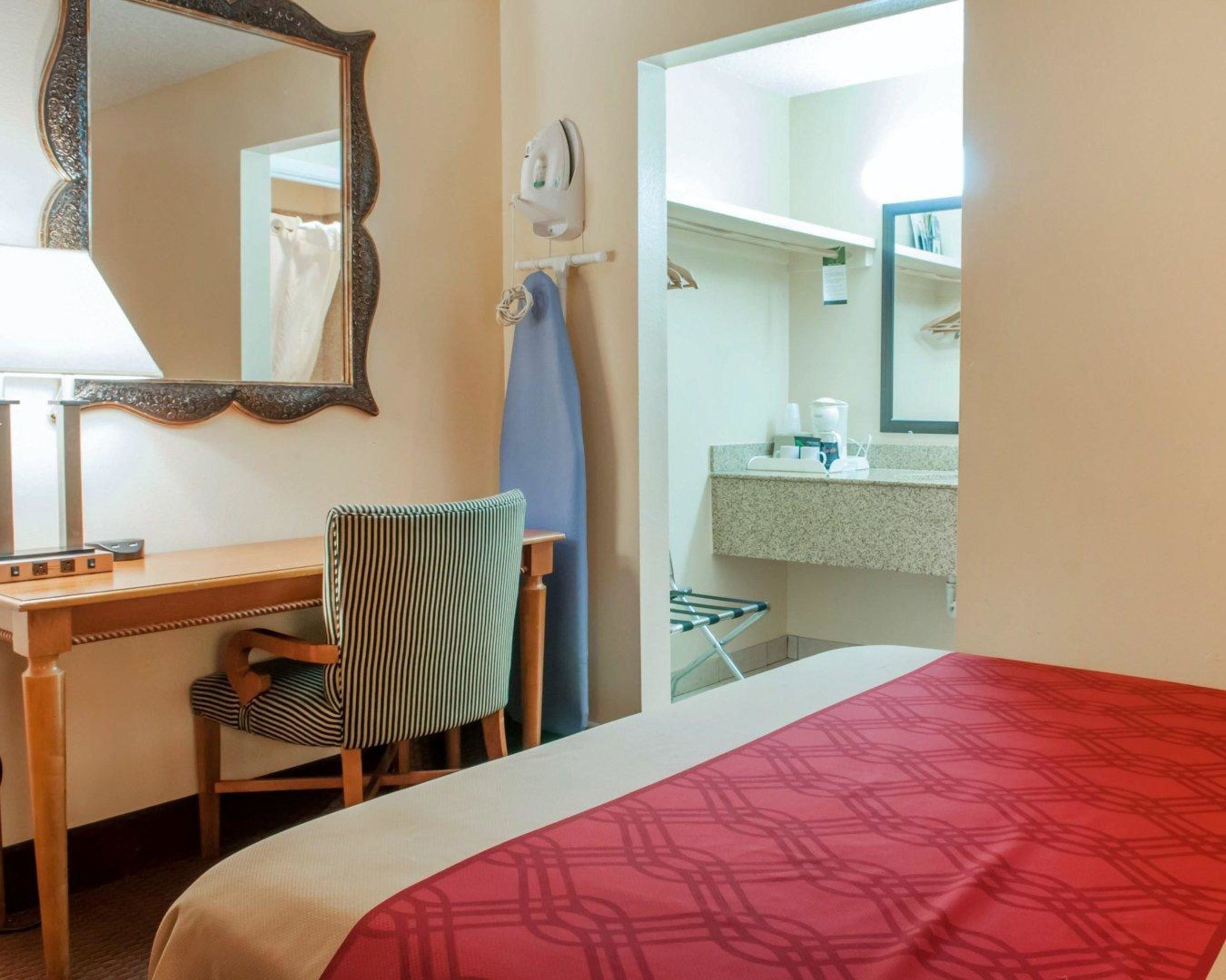 A hotel room with a bed, a desk with a chair, a mirror, a bathroom with a sink, and a shower.