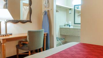 A hotel room with a bed, a desk with a chair, a mirror, a bathroom with a sink, and a shower.
