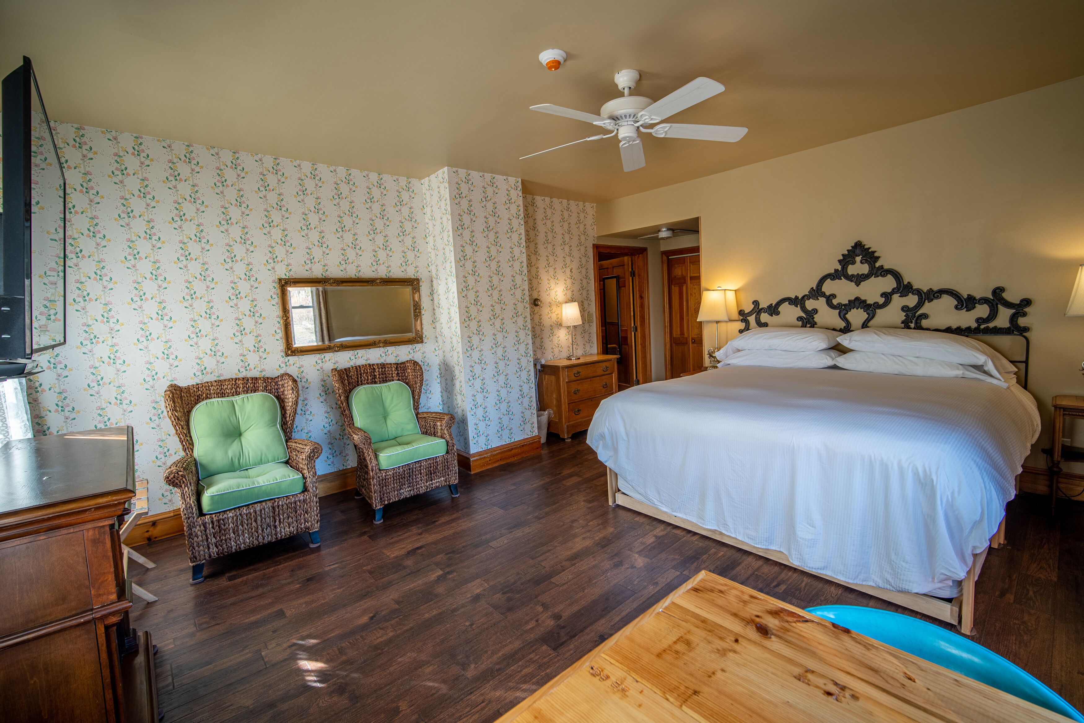 A bedroom with a large bed, two wicker chairs, a wooden desk, a ceiling fan, and a mirror on the wall.