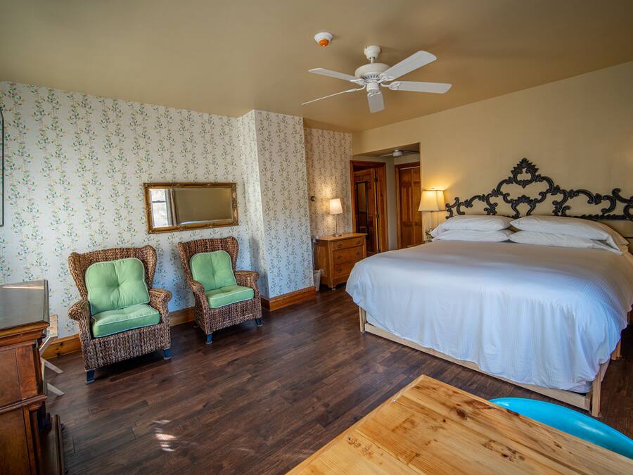 A bedroom with a large bed, two wicker chairs, a wooden desk, a ceiling fan, and a mirror on the wall.