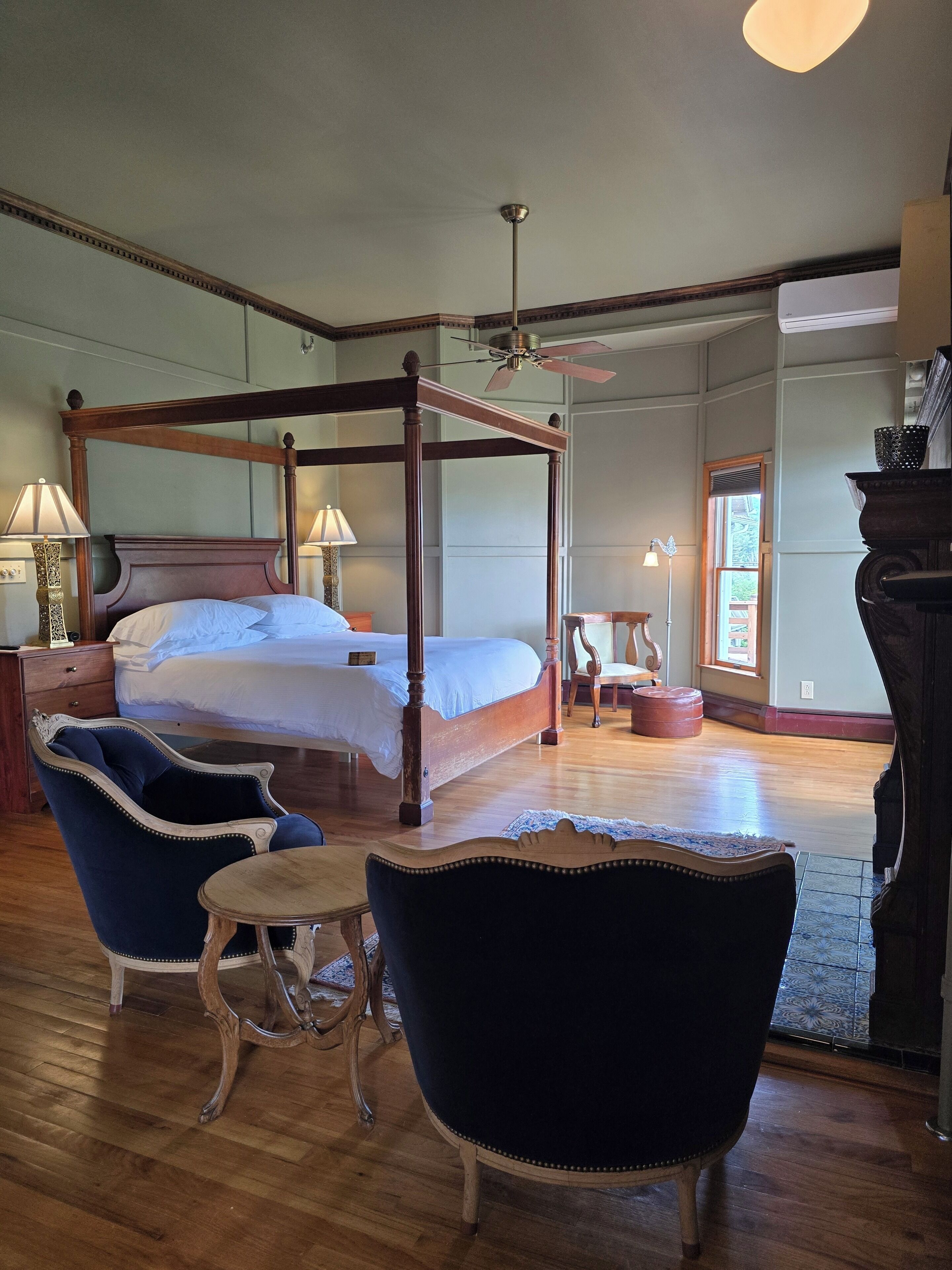 A four-poster bed with a ceiling fan, two upholstered chairs, a small table, and a wooden floor.