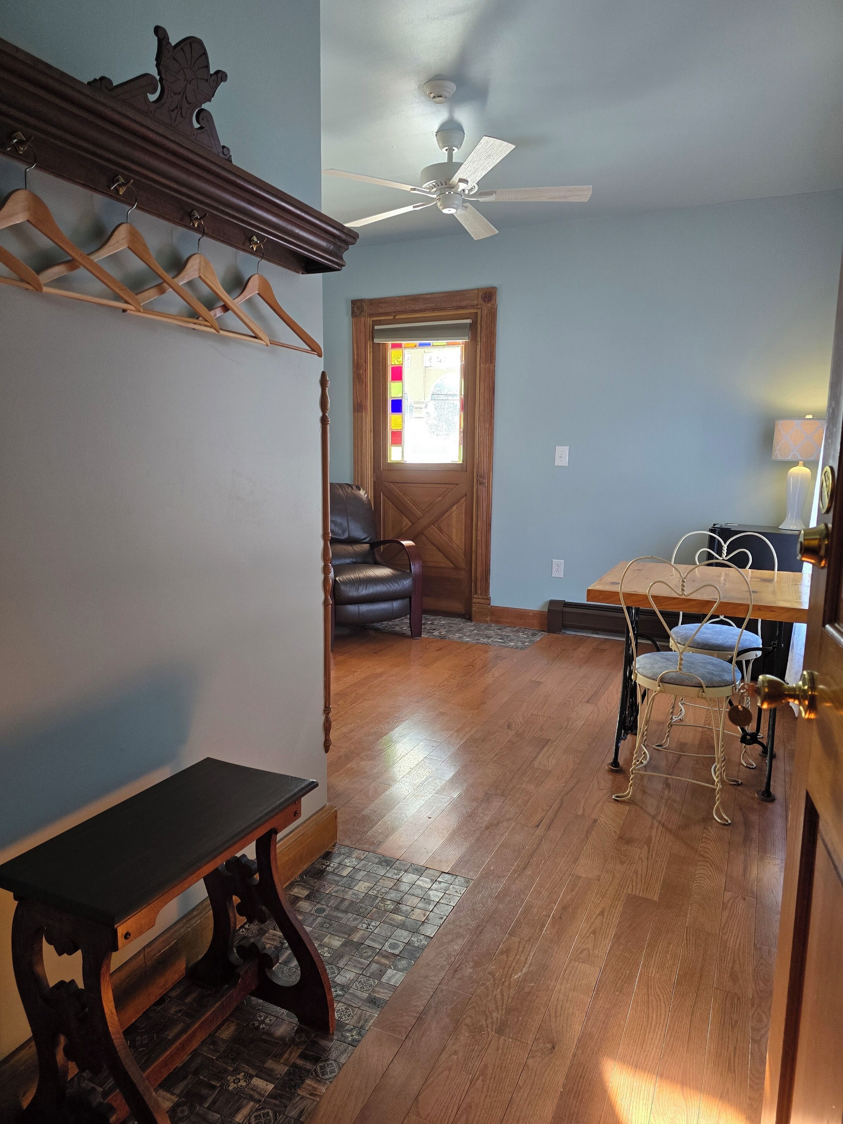 A room with a wooden floor, a ceiling fan, a bench, a table with a heart-shaped chair, and a door with a stained glass window.