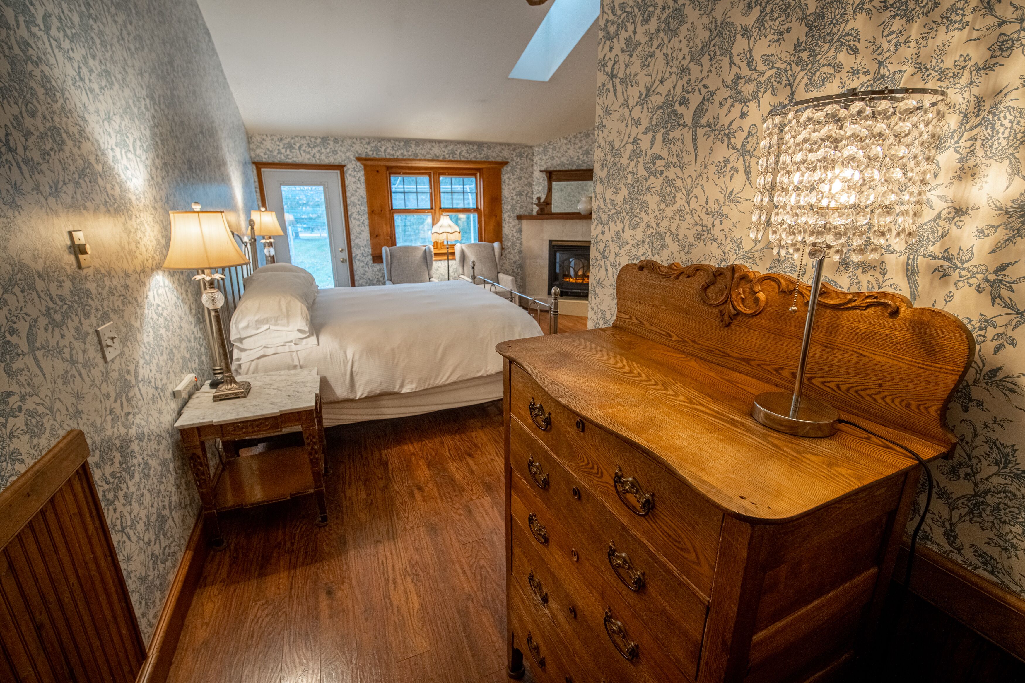 A bedroom with a bed, wooden dresser, two chairs, a nightstand, a lamp, and a fireplace.