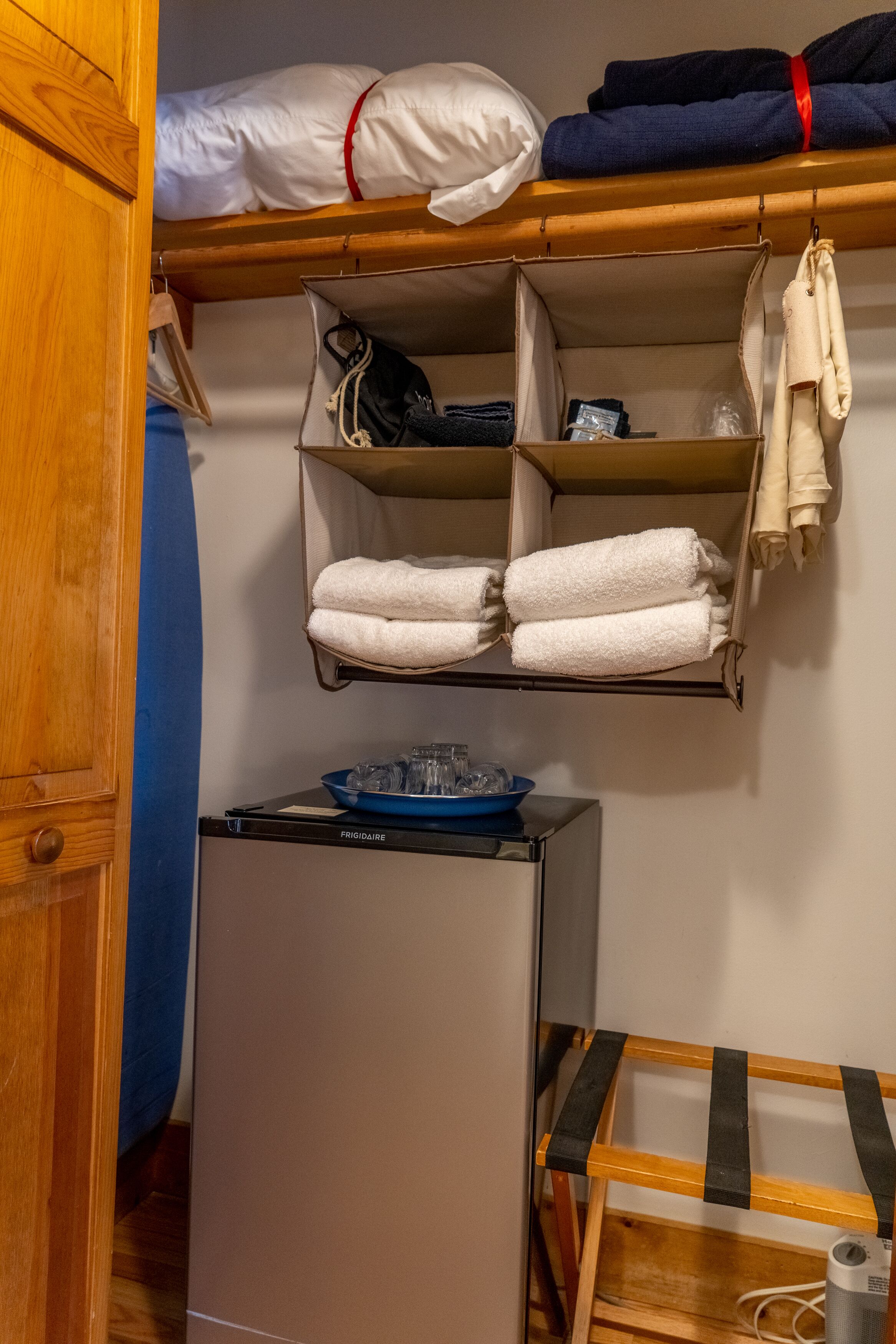 Una habitación compacta con literas, un refrigerador pequeño y un estante con toallas dobladas y una bolsa.