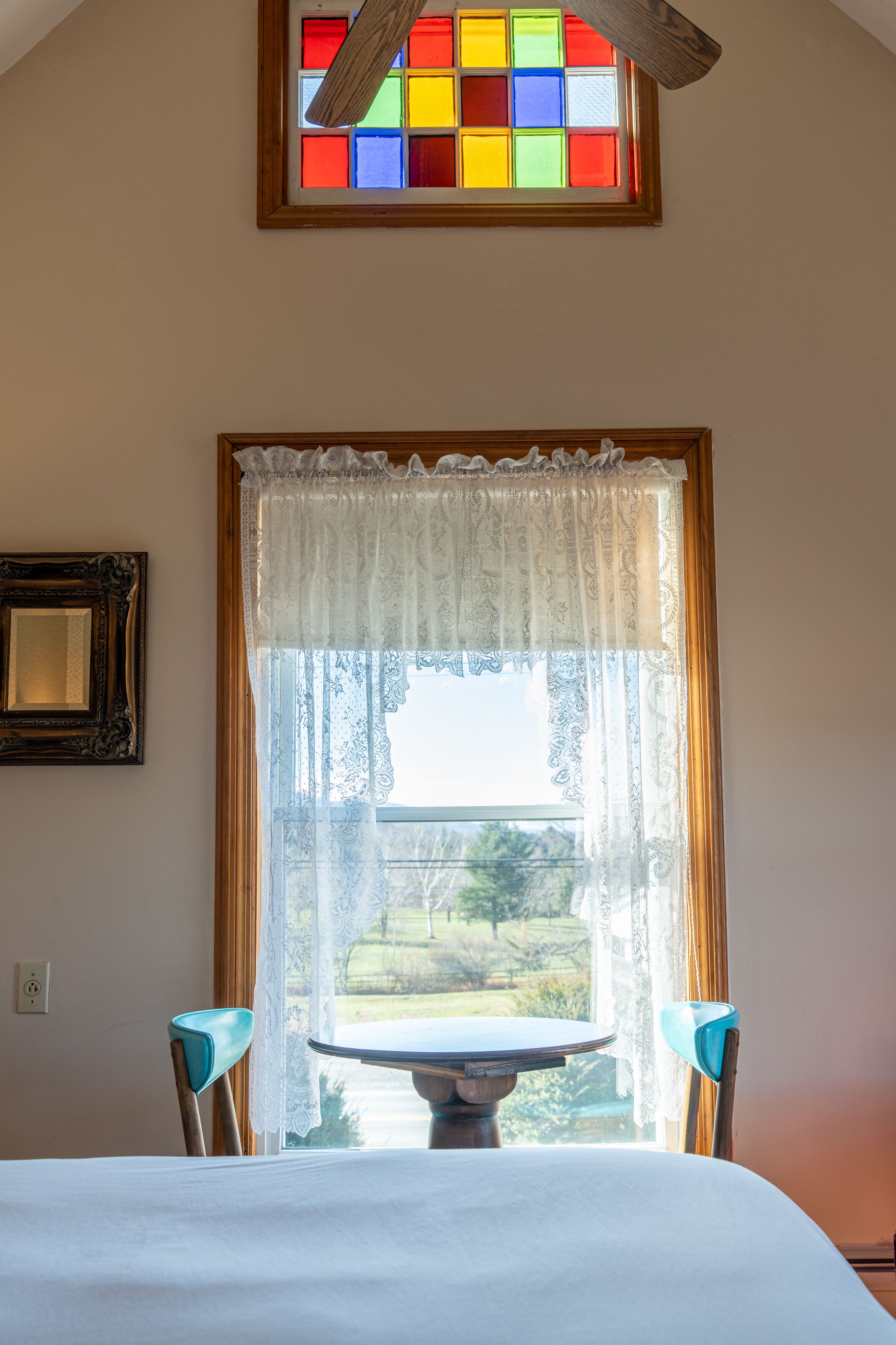 A room with a dining table, chairs, a window with lace curtains, and a stained glass window above.