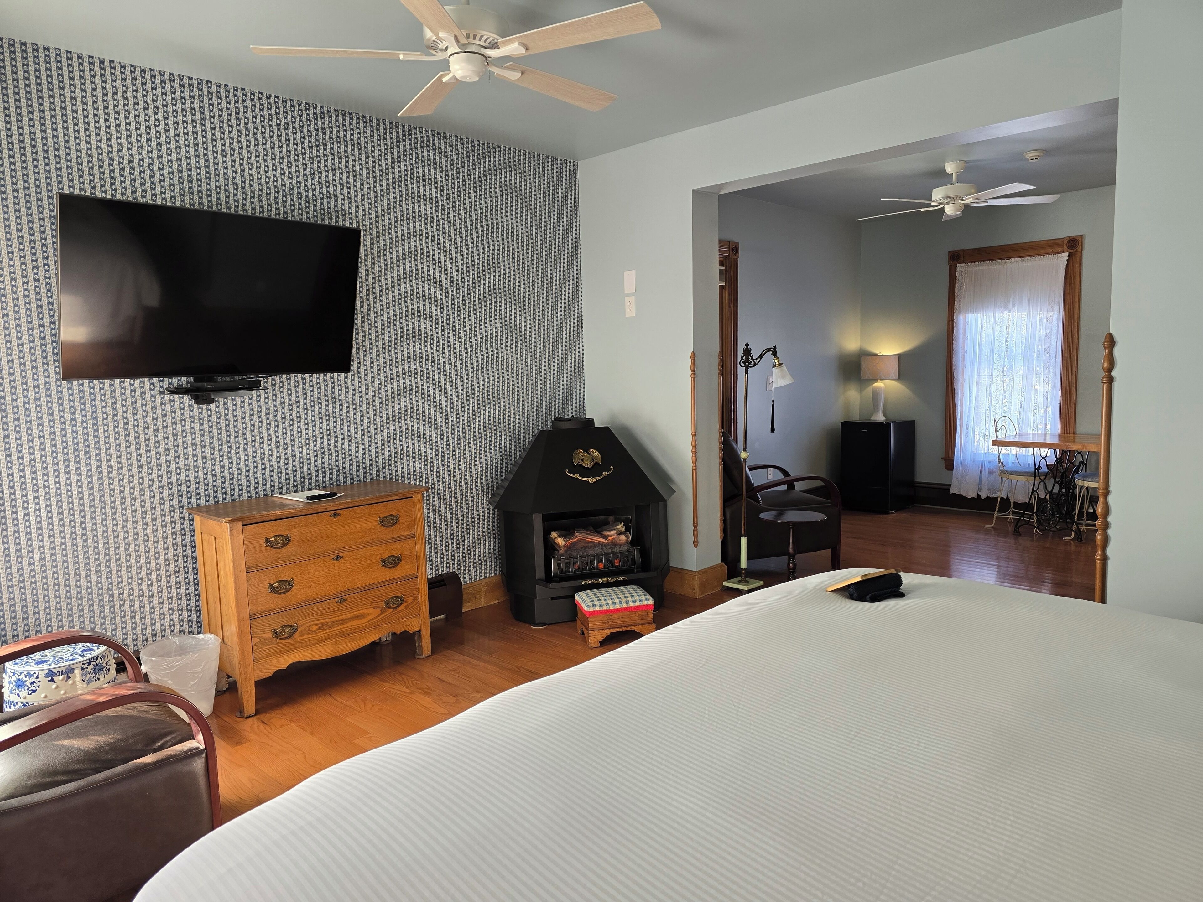 A hotel room with a large bed, a wooden dresser, a fireplace, a television, and a dining area with a table and chairs.