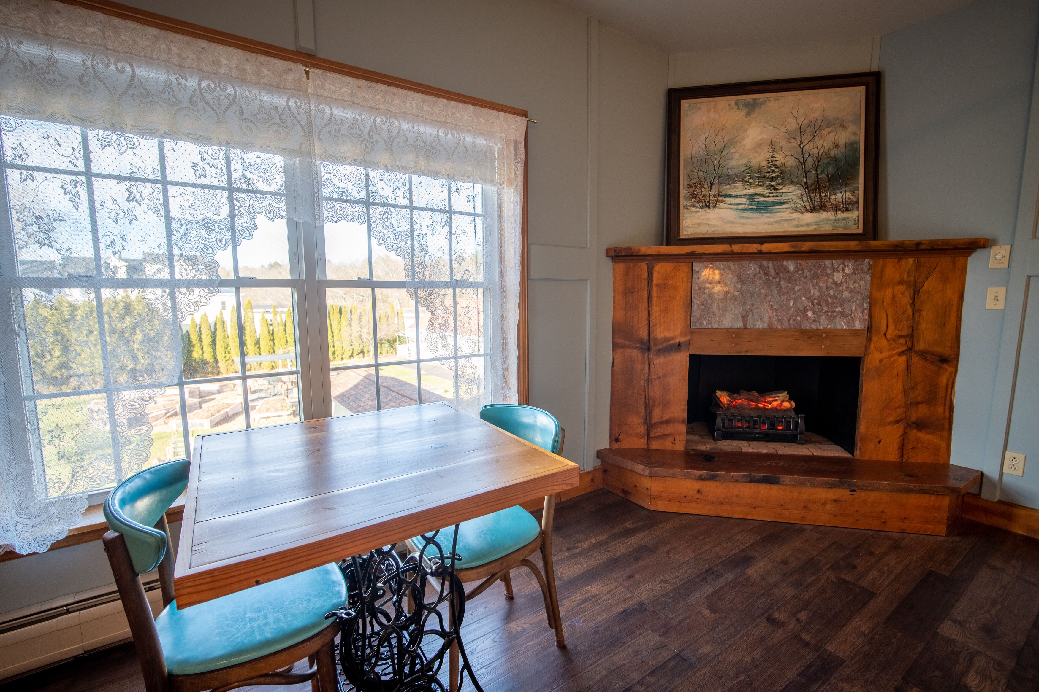 A room with a wooden table and chairs, a fireplace with a painting above it, and a large window with lace curtains.