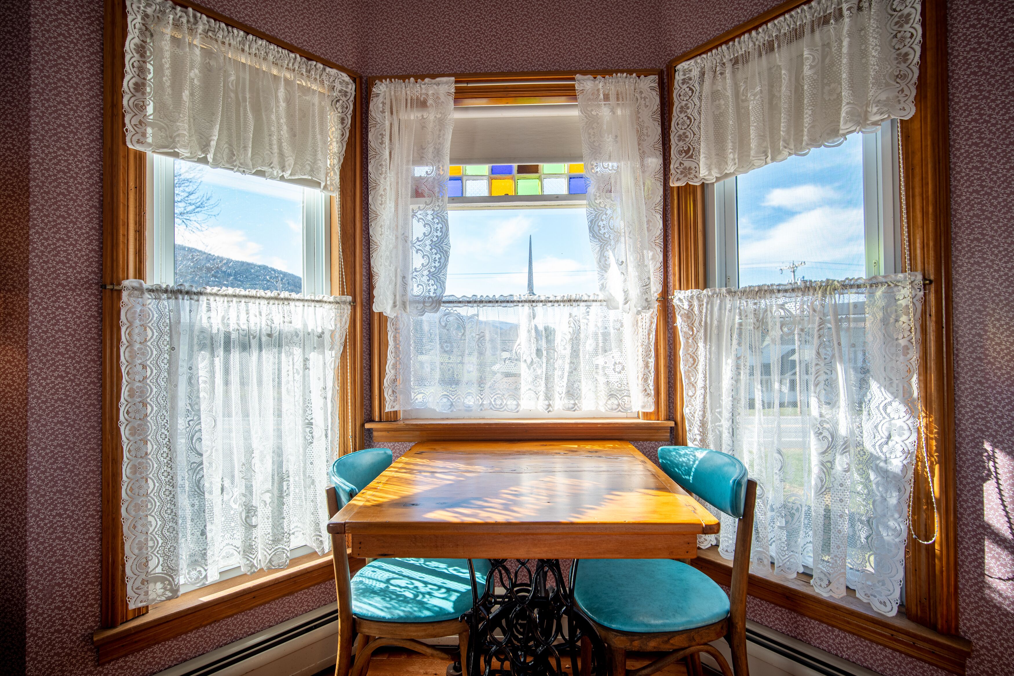 A dining area with a wooden table and four chairs, surrounded by lace curtains and a window with a stained glass view.