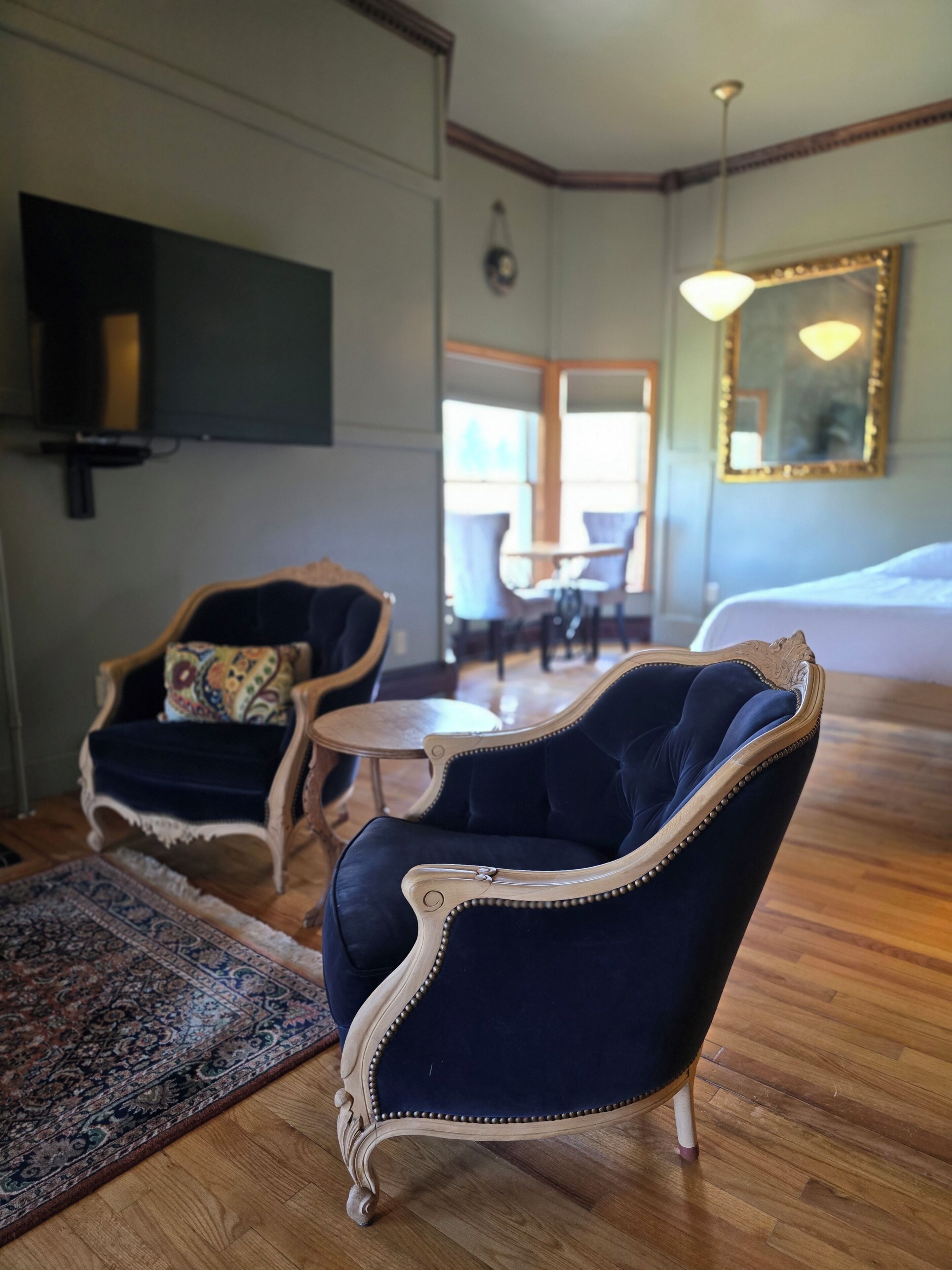 A room with two dark blue upholstered chairs with ornate detailing, a small round table, a patterned rug, a television mounted on the wall, and a large mirror with a decorative frame.