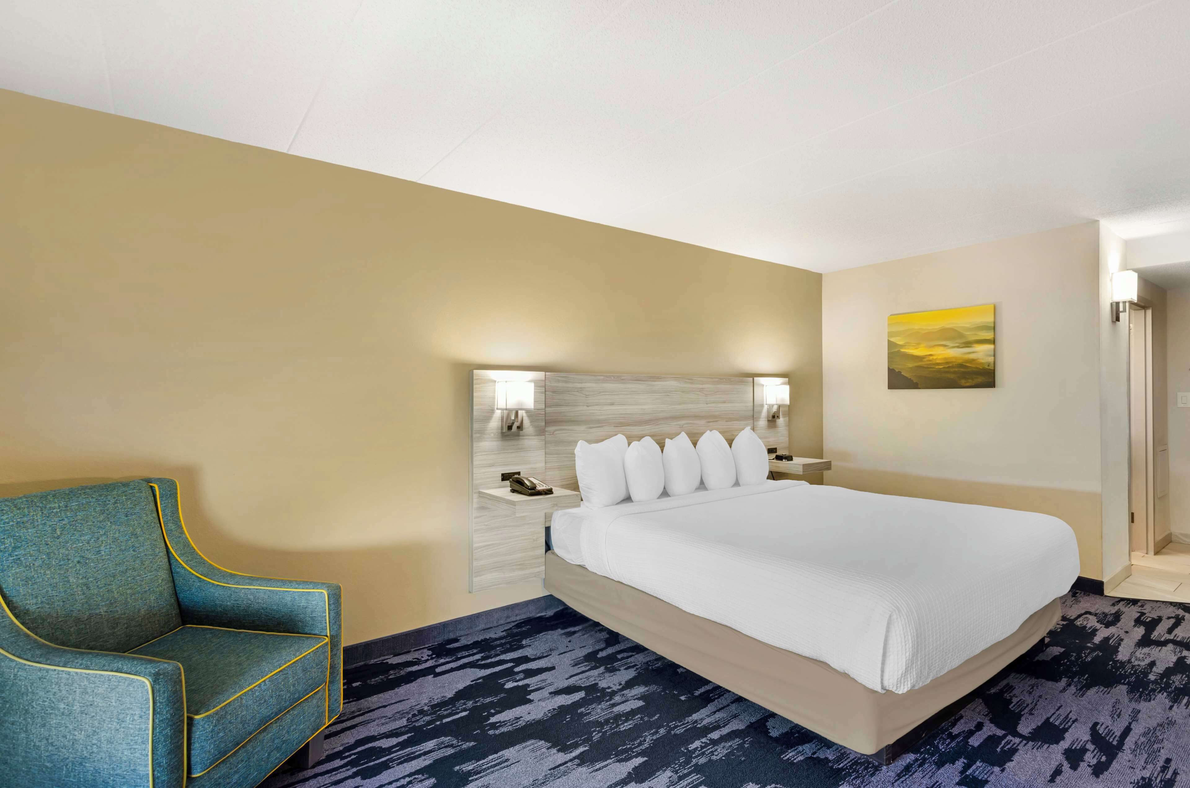 A hotel room with a large bed, a blue armchair, a nightstand with a phone, and a painting on the wall.