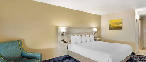A hotel room with a large bed, a blue armchair, a nightstand with a phone, and a painting on the wall.