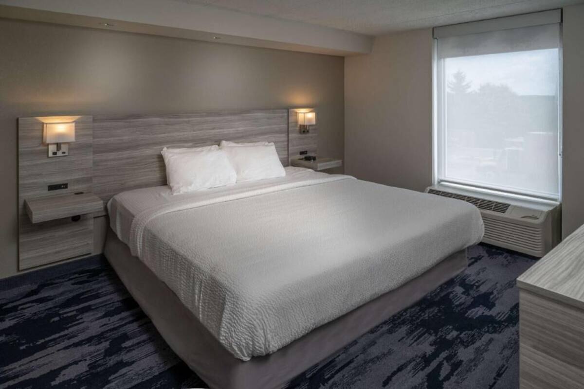A hotel room with a large bed, two bedside tables with lamps, a window with a view, and a built-in air conditioning unit.