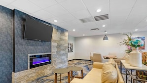 Un vestíbulo de hotel con una televisión de pantalla plana montada en una pared de piedra, una chimenea pequeña, un sofá y una zona de comedor con mesas y sillas.