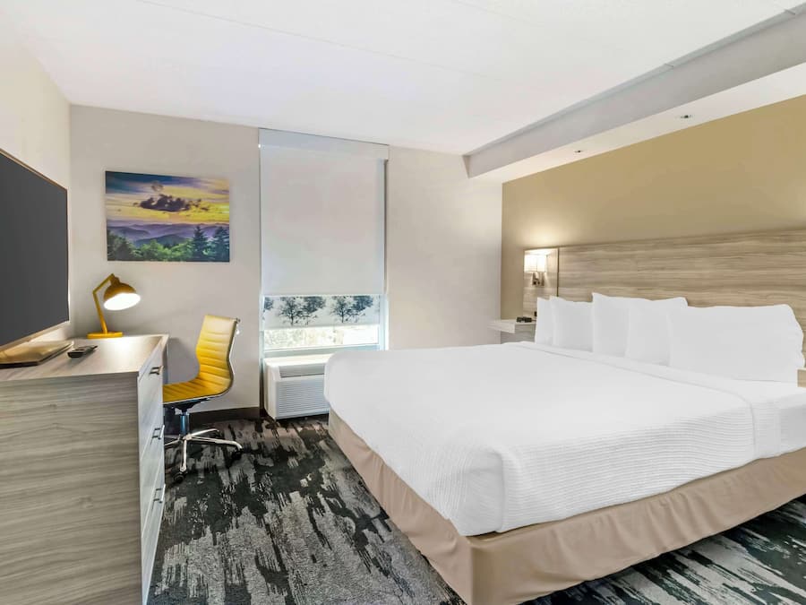 A modern hotel room with a large bed, a desk with a lamp, a chair, and a painting on the wall.